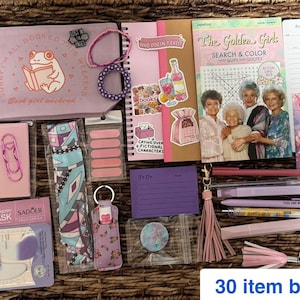 May include: A 30-item stationery and self-care bundle featuring a light purple zippered pouch with a frog illustration, a Golden Girls search and color book, various pens, a lip mask, bookmarks, stickers, a reading log, and a beaded bracelet.