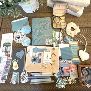 May include: An assortment of stationery and accessories, including a journal, stickers, a small pouch, and a jewelry box. The items are in shades of blue, white, and beige, arranged on a wooden surface. A small white heart-shaped item is also present.