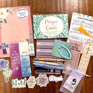 May include: A collection of religious stationery including a pink journal with floral accents, a box of prayer cards, a books of the bible chart, a wooden bookmark with a yellow tassel, pens, stickers, a beaded bracelet, and a small prayer list card.