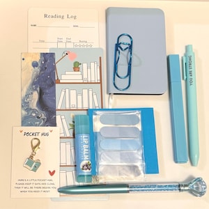 May include: A collection of blue stationery items, including a notebook, pens, sticky notes, and a bookmark. The image also features a lip balm, a paperclip, and a charm with the text "POCKET HUG". The pen has a diamond-shaped top.