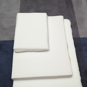 Könnte beinhalten: Drei weiße rechteckige Memory-Schaumstoff-Matratzenauflagen, die übereinander auf einem grauen und blauen gemusterten Teppich gestapelt sind.