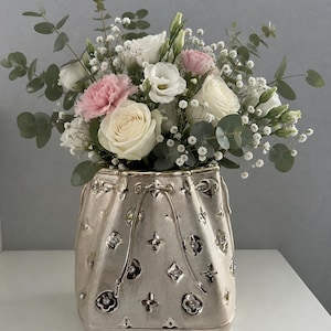 Könnte beinhalten: Eine metallisch champagnerfarbene Keramikvase in Form eines Beutels mit Kordelzug, verziert mit geprägten Blumen- und geometrischen Mustern sowie eingebetteten klaren Kristallen, gefüllt mit weißen Rosen, rosa Nelken und Eukalyptusblättern.
