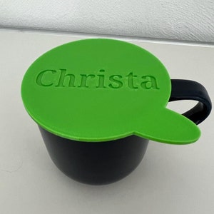 Könnte beinhalten: Eine schwarze Kaffeetasse mit einem grünen Deckel. Der Deckel hat die Form einer Sprechblase und trägt den Namen "Christa" als Prägung. Die Tasse hat einen schwarzen Henkel.