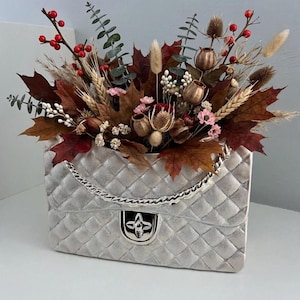 Könnte beinhalten: Eine silberne gesteppte Handtasche mit einer metallischen Kettenschulterriemen und einem silbernen Verschluss, gefüllt mit einem Arrangement aus getrocknetem Herbstlaub, braunen Ahornblättern, roten Beeren, Weizenähren und kleinen rosa Blüten.