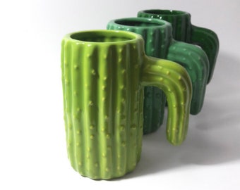May include: Three green ceramic mugs shaped like cacti with textured surfaces. The mugs have handles that are also shaped like cacti.