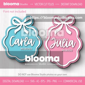 May include: Two layered cut files with the names "Carla" and "Julia". The "Carla" design is light blue with white accents, and the "Julia" design is pink with white accents. Both have a bow at the top and the names are in cursive. The text "blooma Studio" is also present.