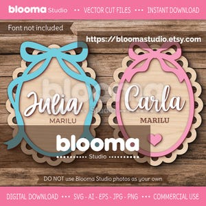 以下が含まれることがあります： 「Julia」と「Carla」の名前が、リボンが付いた装飾的な楕円形のフレームに入った木製のカットファイル。「Marilu」は各名前の下に書かれています。「Julia」のデザインは青いリボンで、「Carla」のデザインはピンクのリボンとハートが付いています。Blooma Studioのロゴが下にあります。