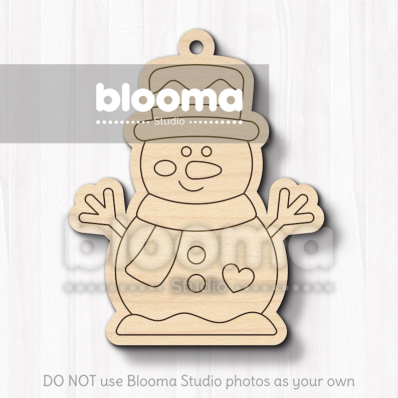 May include: Wooden snowman ornament with a hat, scarf, and heart detail. The snowman has a smiling face and outstretched arms. The ornament has a hole at the top for hanging. The text "blooma studio" is visible on the ornament.