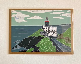 Op de afbeelding: Een kaart met een kleurrijke illustratie van een vuurtoren op een klif met uitzicht op de zee. De kaart heeft een groen, blauw en wit kleurenschema en bevat de tekst "Sorrell Reilly".