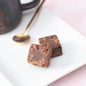 May include: Two dark brown sugar cubes with red flecks on a white plate. The sugar cubes are on a white plate with a pink background.