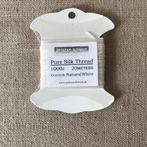May include: A white spool of 1000d pure silk thread in a plastic dispenser. The label reads "Pure Silk Thread 1000d 20 meters Colour: Natural White www.spitzen-borchen.de"