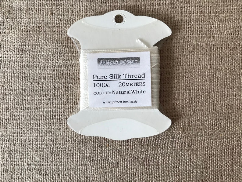 May include: A white spool of 1000d pure silk thread in a plastic dispenser. The label reads "Pure Silk Thread 1000d 20 meters Colour: Natural White www.spitzen-borchen.de"