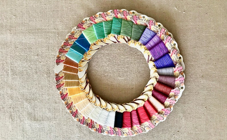 May include: A circular display of 24 spools of embroidery floss in various colors, including green, blue, brown, yellow, white, black, red, purple, and gray. The spools are arranged in a rainbow pattern and are labeled "Sea Lace".