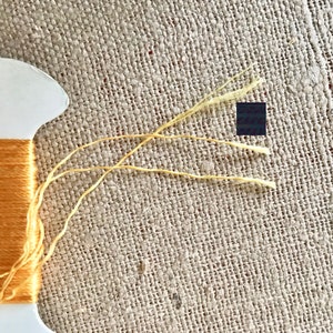 May include: A spool of orange embroidery floss with three strands of yellow floss extending from the spool.