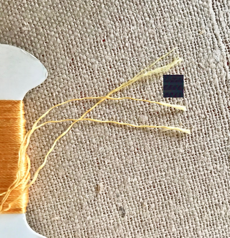 May include: A spool of orange embroidery floss with three strands of yellow floss extending from the spool.
