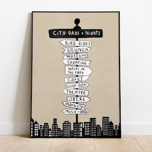 May include: A framed poster with a beige backdrop, displaying a black signpost with arrows indicating city activities. Text includes "City Days & Nights", "Bike Rides", "Brunch", "Museums", and "Shopping". A black cityscape silhouette is at the bottom.