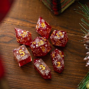 May include: Set of seven red and gold dice with white snowflake designs. The dice are made of clear resin with a glittery finish.
