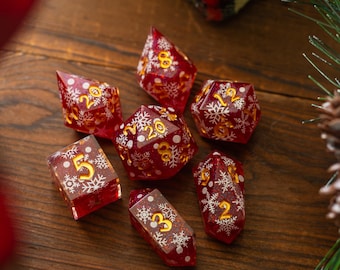 May include: Set of seven red and gold dice with white snowflake designs. The dice are made of clear resin with a glittery finish.