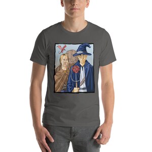 May include: A dark gray t-shirt with a graphic print of a wizard and a woman standing in front of a castle. The wizard is wearing a blue robe and a pointed hat. The woman is wearing a brown dress. A red dragon is flying in the sky above them.