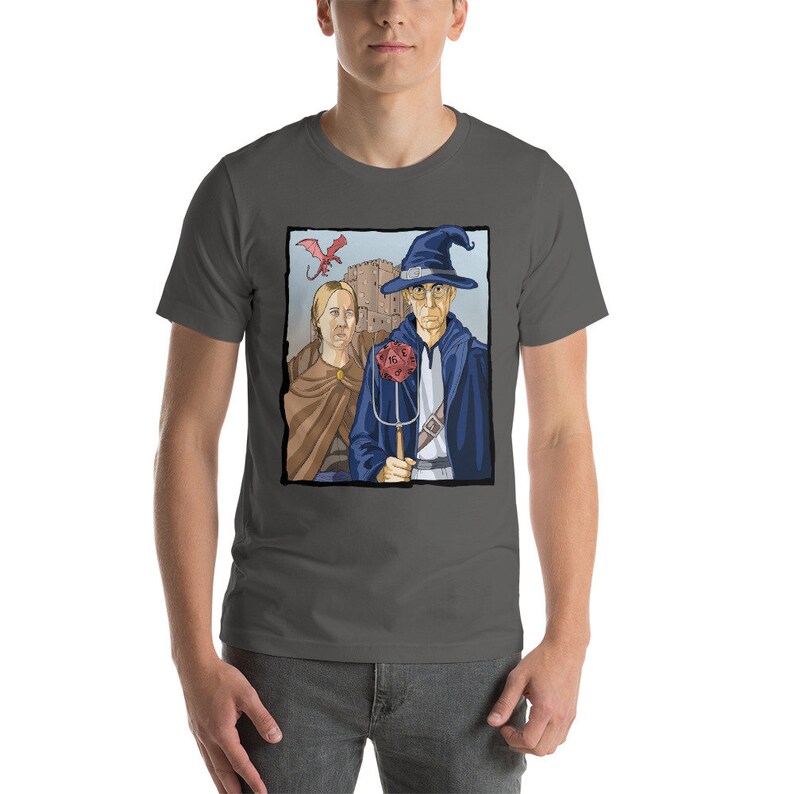 May include: A dark gray t-shirt with a graphic print of a wizard and a woman standing in front of a castle. The wizard is wearing a blue robe and a pointed hat. The woman is wearing a brown dress. A red dragon is flying in the sky above them.
