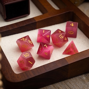 May include: A set of seven pink and gold dice with numbers 1, 4, 7, 19, 20, 30, and 60. The dice are on a wooden tray with a white cloth lining.