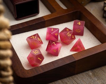 May include: A set of seven pink and gold dice with numbers 1, 4, 7, 19, 20, 30, and 60. The dice are on a wooden tray with a white cloth lining.