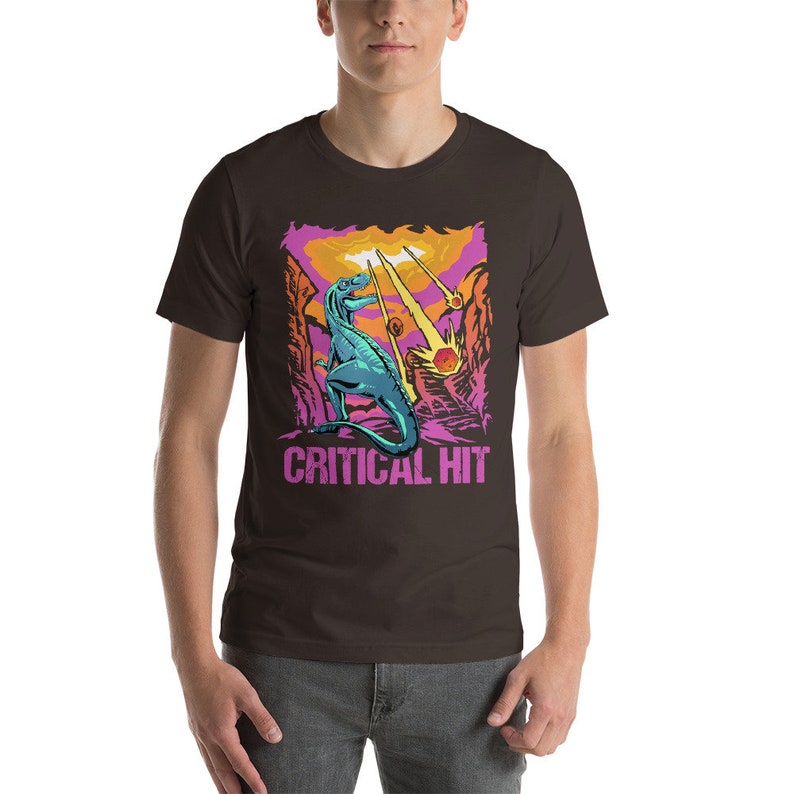 May include: A brown t-shirt with a graphic of a blue dinosaur standing in a canyon with falling stars and the text "CRITICAL HIT".