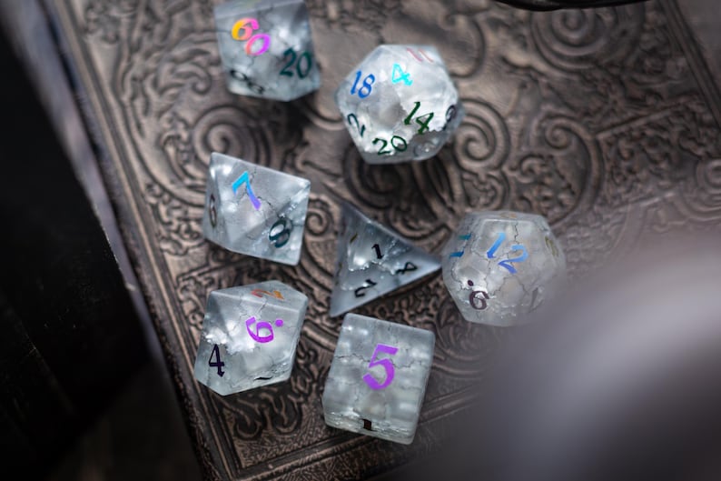 K&ouml;nnte beinhalten: Ein Satz von sieben schimmernden W&uuml;rfeln mit wei&szlig;en und blauen Zahlen. Die W&uuml;rfel sind aus klarem Kunststoff und haben eine matte Oberfl&auml;che. Die W&uuml;rfel liegen auf einer dunkelbraunen Oberfl&auml;che mit einem dekorativen Muster.