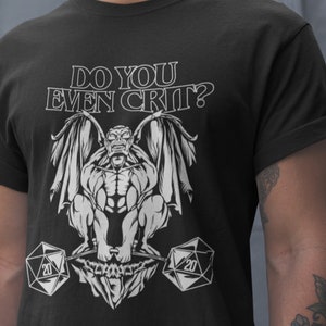以下が含まれることがあります： 黒いTシャツ。白いグラフィックは、D20サイコロを重りとしてバーベルを持ち上げるガーゴイル。テキスト「DO YOU EVEN CRIT？」はガーゴイルの上にあります。
