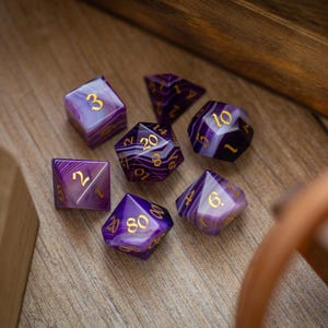 Könnte beinhalten: Sieben violett-weiß marmorierte, polyedrische Würfel mit goldenen Zahlen. Die Würfel, darunter d4, d6, d8, d10, d12 und d20 Formen, liegen auf einer Holzoberfläche, ideal für Tabletop-Spiele.