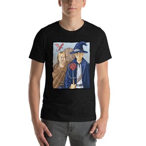 May include: A black t-shirt with a graphic print of a wizard and a woman standing in front of a castle. A red dragon flies above them. The wizard is holding a staff with a large red dice on top. The text "D20" is printed on the shirt.
