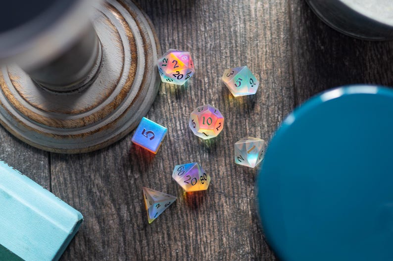 May include: A set of iridescent dice with black numbers on each face. The dice are on a wooden surface with a blue container and a white pedestal.