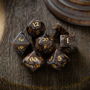 May include: A set of seven black and gold dice with various numbers on each side. The dice are made of a speckled material that resembles stone.