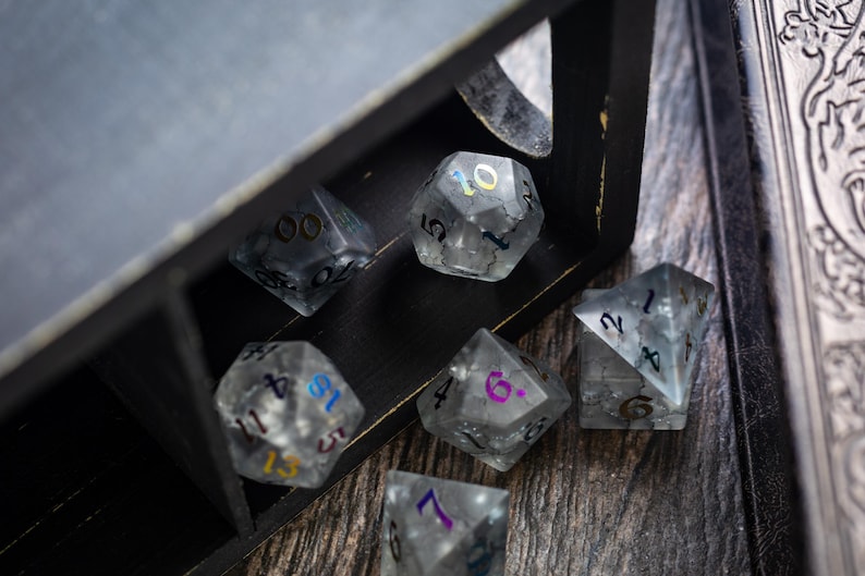 K&ouml;nnte beinhalten: Ein Satz von sechs klaren, schillernden W&uuml;rfeln mit bunten Zahlen. Die W&uuml;rfel befinden sich in einer Holzkiste mit schwarzem Hintergrund.