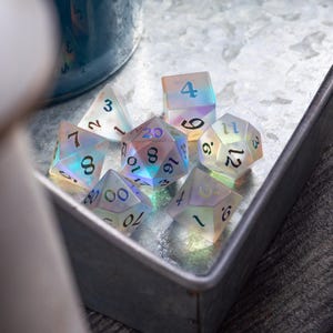 May include: A set of iridescent dice with black numbers. The dice are in a metal tray with a white background. The numbers on the dice are 3, 12, 16, 9, 6, 4, 16, 8, 70, 0000, 10, 8, NL, 3, 20, 3.