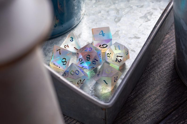 May include: A set of iridescent dice with black numbers. The dice are in a metal tray with a white background. The numbers on the dice are 3, 12, 16, 9, 6, 4, 16, 8, 70, 0000, 10, 8, NL, 3, 20, 3.