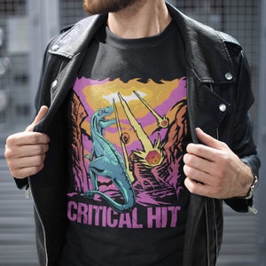 May include: A black t-shirt with a graphic of a dinosaur standing on a rocky landscape with a pink, orange, and yellow sky. The dinosaur is blue and green. The text "CRITICAL HIT" is printed in pink below the dinosaur.