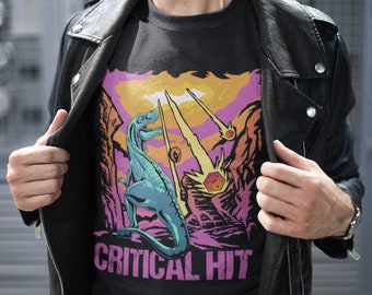 May include: A black t-shirt with a graphic of a dinosaur standing on a rocky landscape with a pink, orange, and yellow sky. The dinosaur is blue and green. The text "CRITICAL HIT" is printed in pink below the dinosaur.