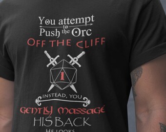 以下が含まれることがあります： 白いグラフィックデザインが施された黒いTシャツ。デザインには、1の目のあるサイコロ、交差した2本の剣、そして「You attempt to Push the Orc OFF THE CLIFF. Instead, you gently massage HIS BACK. He looks UNCOMFORTABLE.」というテキストが描かれています。