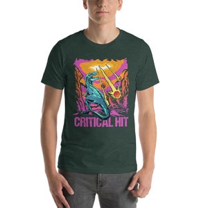 May include: A green t-shirt with a graphic of a blue dinosaur standing on a pink and orange landscape. The dinosaur is facing the left side of the image. The text "CRITICAL HIT" is printed in pink below the dinosaur.