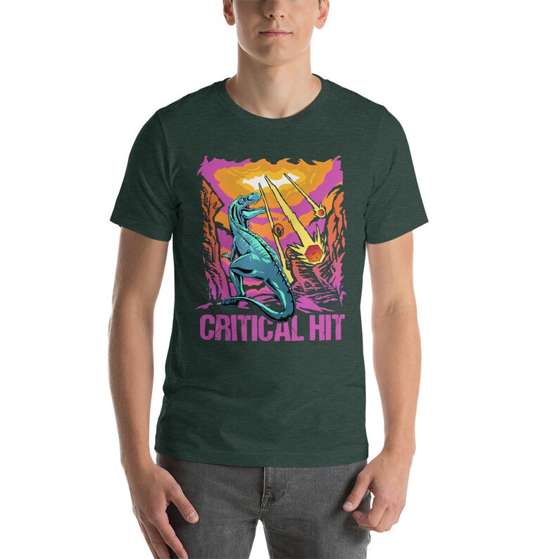 May include: A green t-shirt with a graphic of a blue dinosaur standing on a pink and orange landscape. The dinosaur is facing the left side of the image. The text "CRITICAL HIT" is printed in pink below the dinosaur.