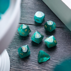 May include: A set of teal green dice with gold numbers. The dice are on a dark wood surface.