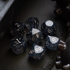 May include: A set of seven polyhedral dice in shades of dark blue and black. The dice have silver numbering and a speckled, glittery appearance. The dice are arranged on a dark surface.