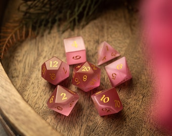 May include: A set of seven pink and white dice with gold numbers. The dice are on a wooden surface.