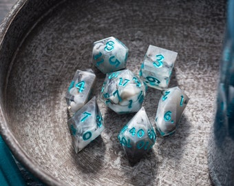 May include: A set of seven polyhedral dice with a marbled gray and white design, teal numbering, and a stone-like interior. The dice are displayed in a rustic metal bowl, with a teal accent visible on the side.