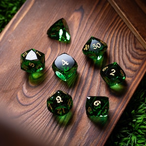 May include: Seven green and gold polyhedral dice are arranged on a wooden surface. The dice are transparent with gold numbering. The wooden surface has a natural wood grain pattern. The dice are for tabletop games.
