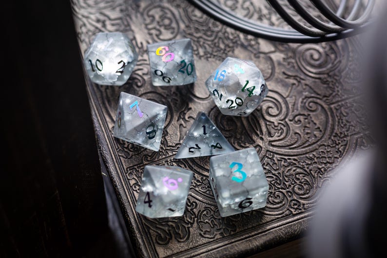 K&ouml;nnte beinhalten: Ein Satz von sechs klaren, schillernden W&uuml;rfeln mit schwarzen und farbigen Zahlen. Die W&uuml;rfel liegen auf einer dunklen, gemusterten Oberfl&auml;che.