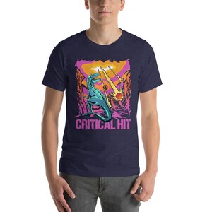 May include: A navy blue t-shirt with a graphic of a green dinosaur standing on a pink and orange landscape with falling stars. The text "CRITICAL HIT" is printed below the dinosaur.