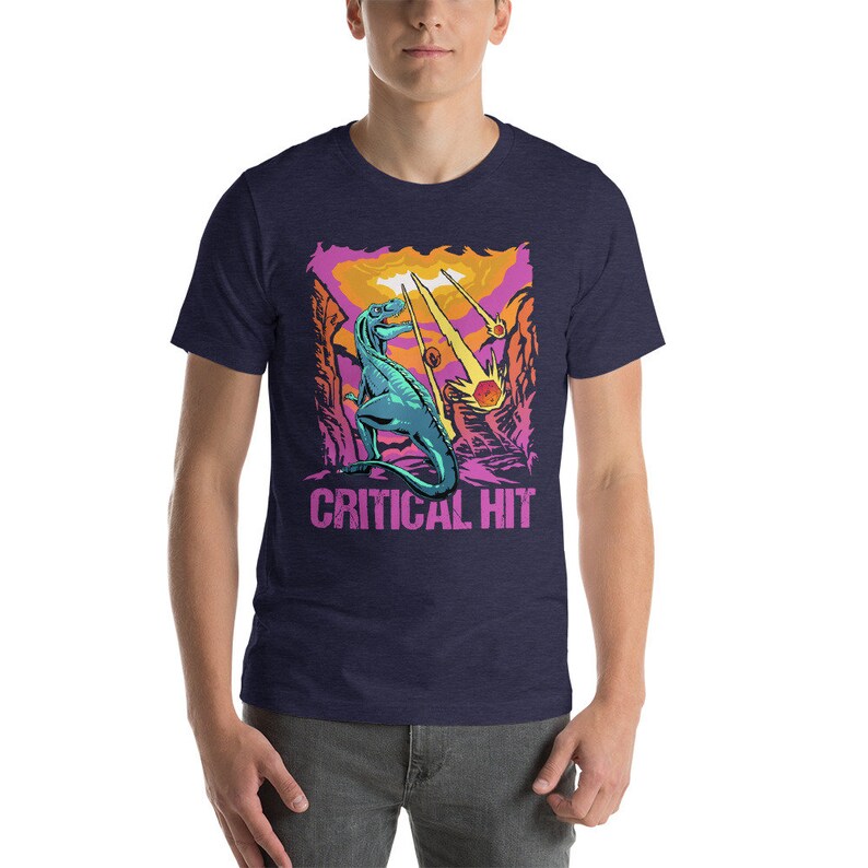 May include: A navy blue t-shirt with a graphic of a green dinosaur standing on a pink and orange landscape with falling stars. The text "CRITICAL HIT" is printed below the dinosaur.