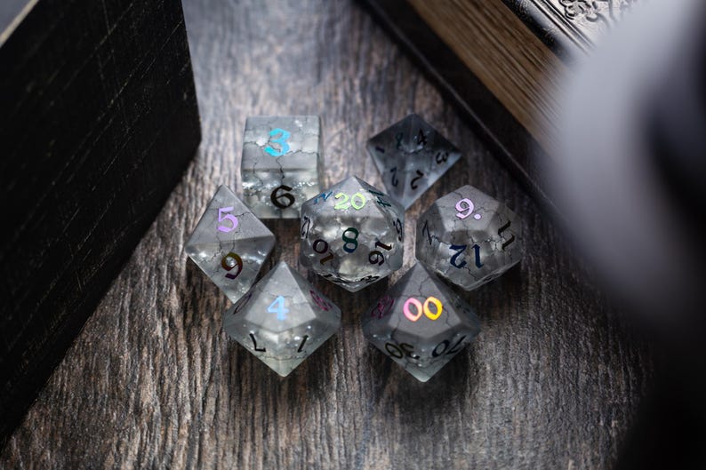 K&ouml;nnte beinhalten: Ein Satz von acht klaren, schillernden W&uuml;rfeln mit schwarzen und bunten Zahlen. Die W&uuml;rfel liegen auf einer dunklen Holzoberfl&auml;che.